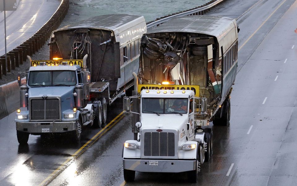 Two damaged train cars are carried on flatbed trailers from the scene of Monday's fatal Amtrak train crash onto Interstate 5 at DuPont. The investigation and cleanup continued throughout Tuesday. — Photograph: Elaine Thompson/The Associated Press.