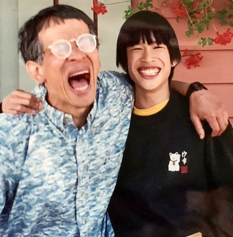 A man in blue shirt and glasses laughs, his arm around a man in a black sweatshirt. 