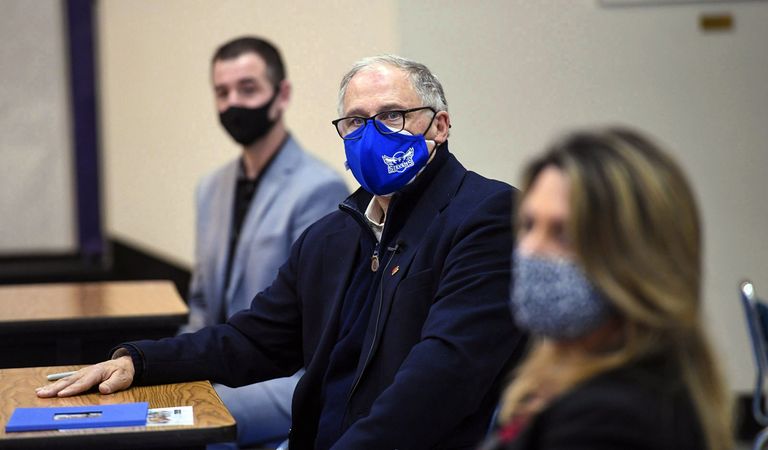 Gov Inslee in a dark blue jacket and blue mask seated at a small wooden desk. Masked people sit on either side of him in the background and foreground. 