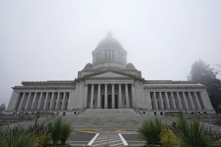 The Washington State Capitol building is partially covered in fog, seen across a parking lot leading up to the building’s light gray steps. 