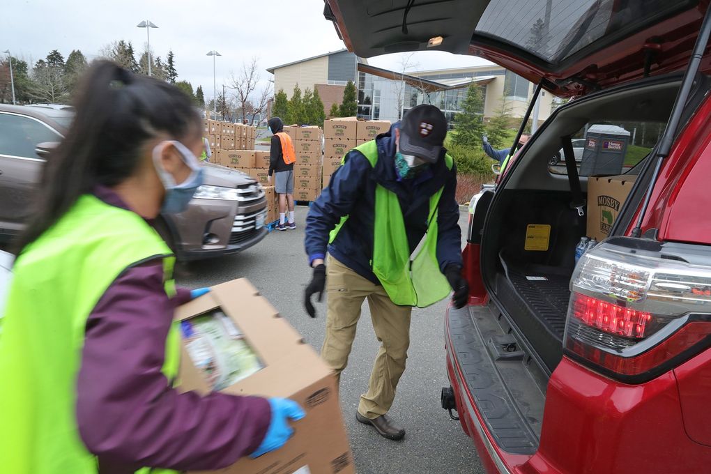 In a parking lot on a cloudy day, two masked people in neon vests walk a box of food toward the open trunk of a red car. 
