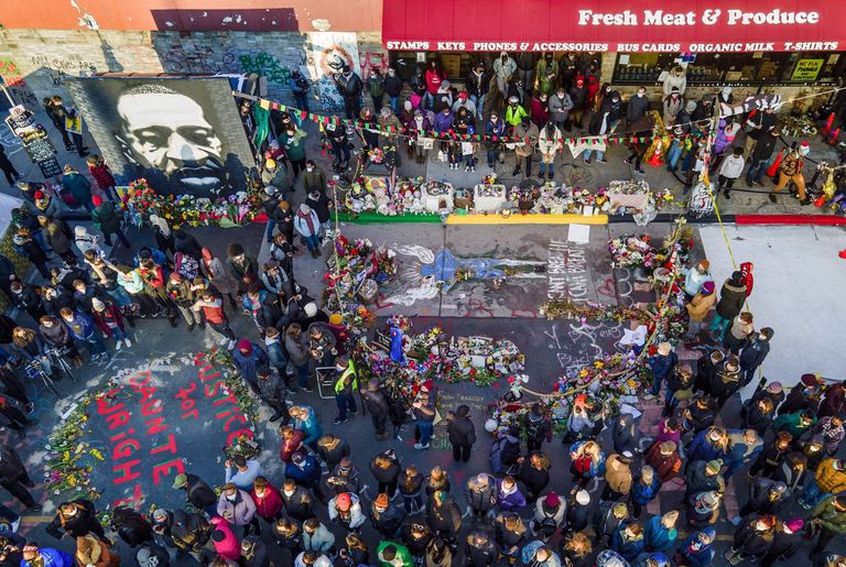 On a street seen from above, a crowd gathers alongside signs, flowers, and murals of George Floyd as the verdict was announced. 