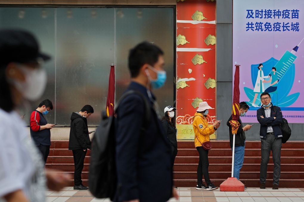 People wearing face masks to help curb the spread of the coronavirus walk by masked residents lining up for COVID-19 vaccine at a vaccination site with a board displaying the slogan, “Timely vaccination to build the Great Wall of Immunity together” in Beijing, Wednesday, April 21, 2021. (AP Photo/Andy Wong)