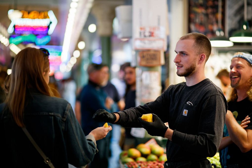 Sam Kallaway, sales lead at Sosio’s Fruit & Produce, center, gives samples of fruit at Pike Place Market in Seattle on Thursday. A new indoor mask mandate goes into effect Monday statewide, reflecting deepening concerns about the delta variant. just as Seattle tourism starts to rebound. (Erika Schultz / The Seattle Times)