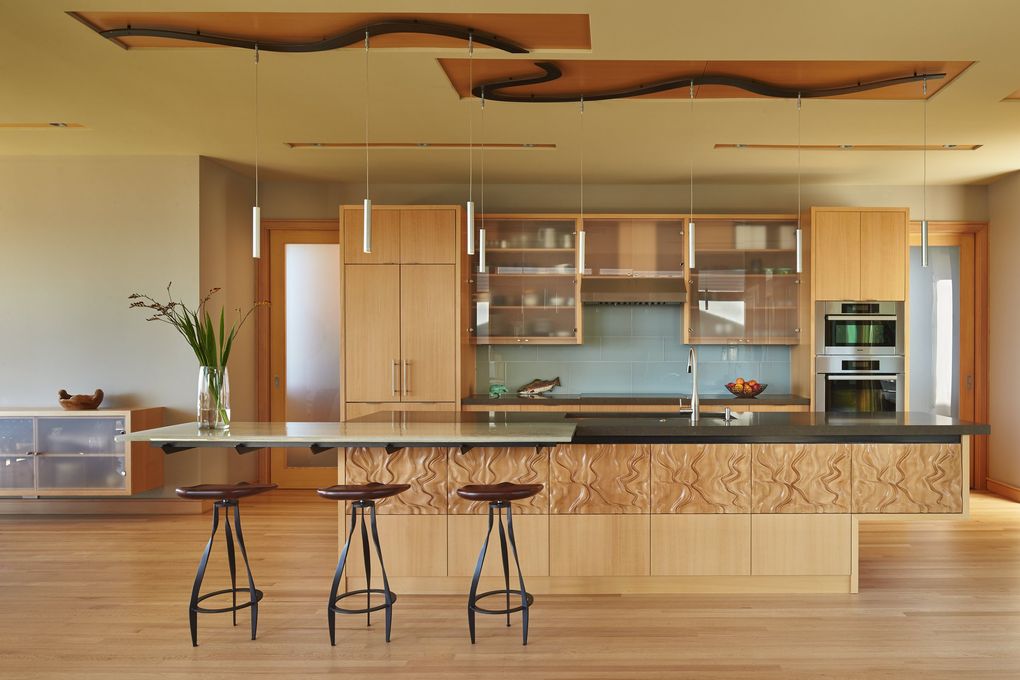 In the brilliantly luminous kitchen of The Elliott Bay House, beech cabinets showcase a custom CNC-milled pattern freehand-sketched by architect Nils Finne; he calls it “imaginary landscape”: river valleys with mountain topography between them. Three sets of free-form, sensuously curved steel lighting bars, drawn “all in one piece” by Finne and crafted by Landbridge Lighting, are set in recessed beech panels in the ceiling to unite the kitchen and adjacent dining area. (Benjamin Benschneider / The Seattle Times, 2016)